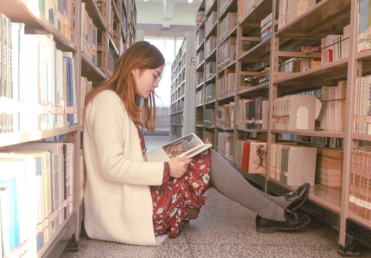 Asian woman sitting and reading book in library aisle, surrounded by bookshelves.