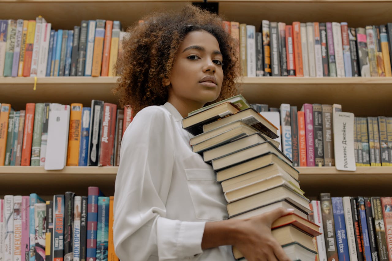 Confident young woman holds stack of books in a library setting.