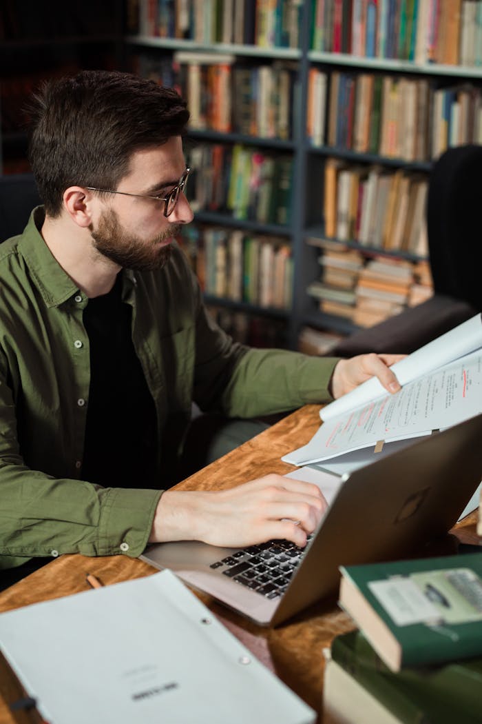 A man studying in a library, working on a laptop and reading documents.